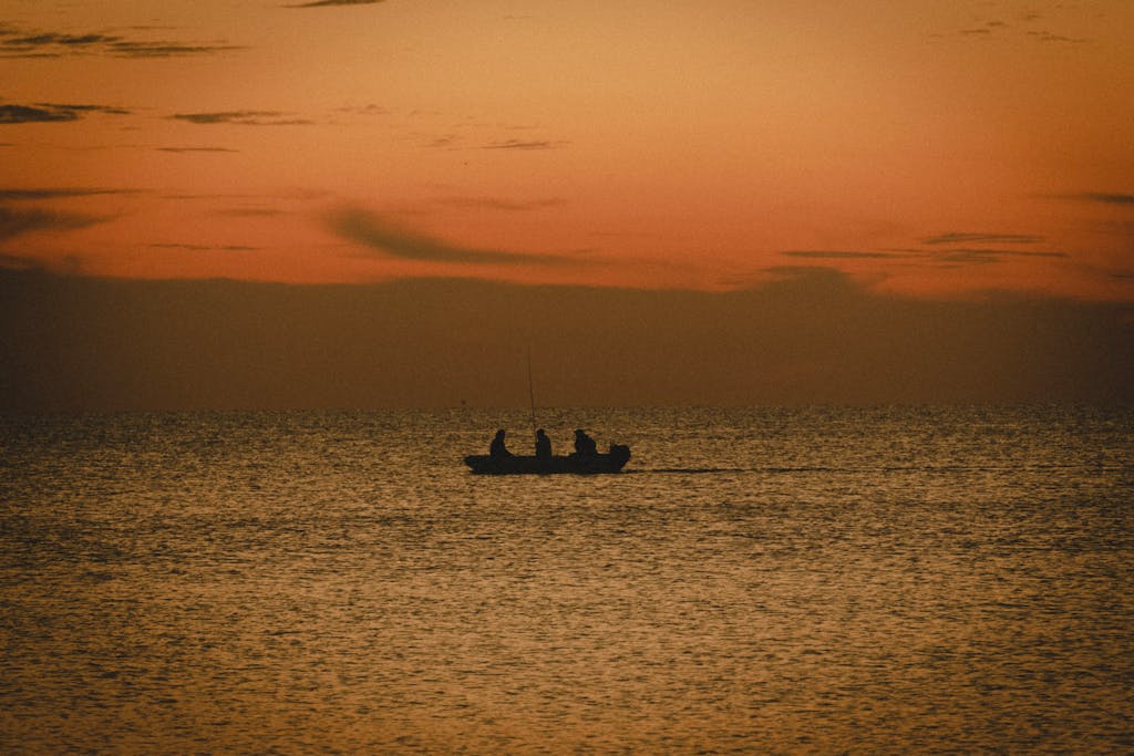 A tranquil scene of a boat with fishermen silhouetted against a stunning orange sunset over the sea.