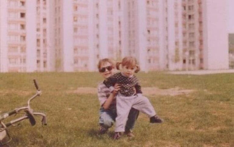 A faded childhood photograph of Nina being held by another child outside apartment buildings.