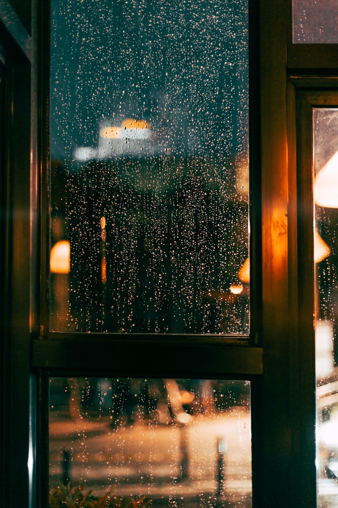 Rain-drenched window capturing urban night lights in a cozy ambiance.