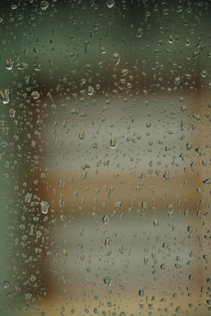Raindrops trickle down a window pane, capturing a blurred view outside, evoking a sense of calmness.
